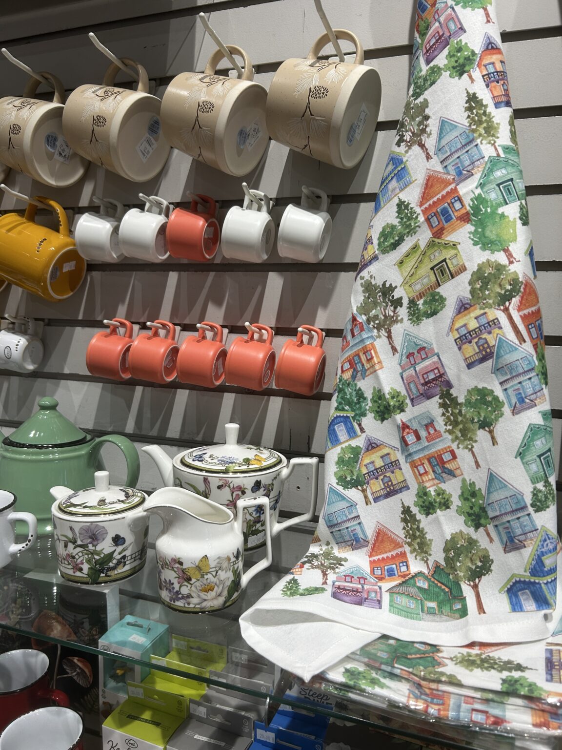 Display table layered with colorful patterned tea towels depicting a European village scene alongside red mugs.