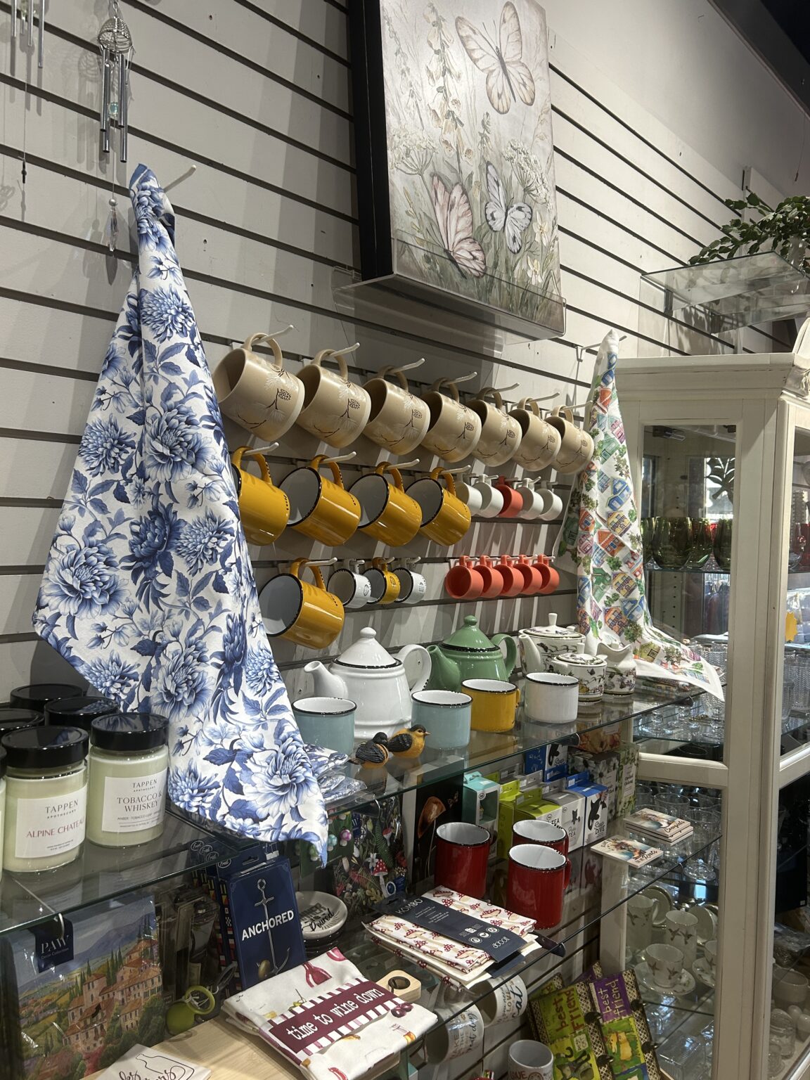 Hanging white tea towels with blue botanical prints draped over a display rack of ceramic mugs.